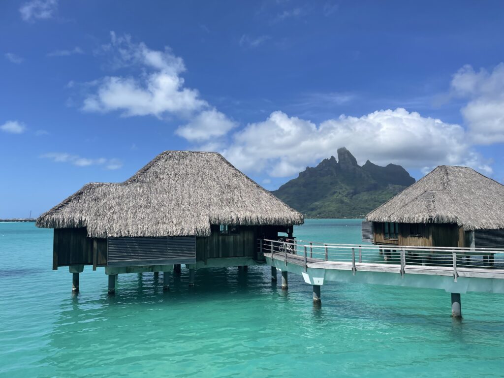 a huts on stilts in the water with Bora Bora in the background